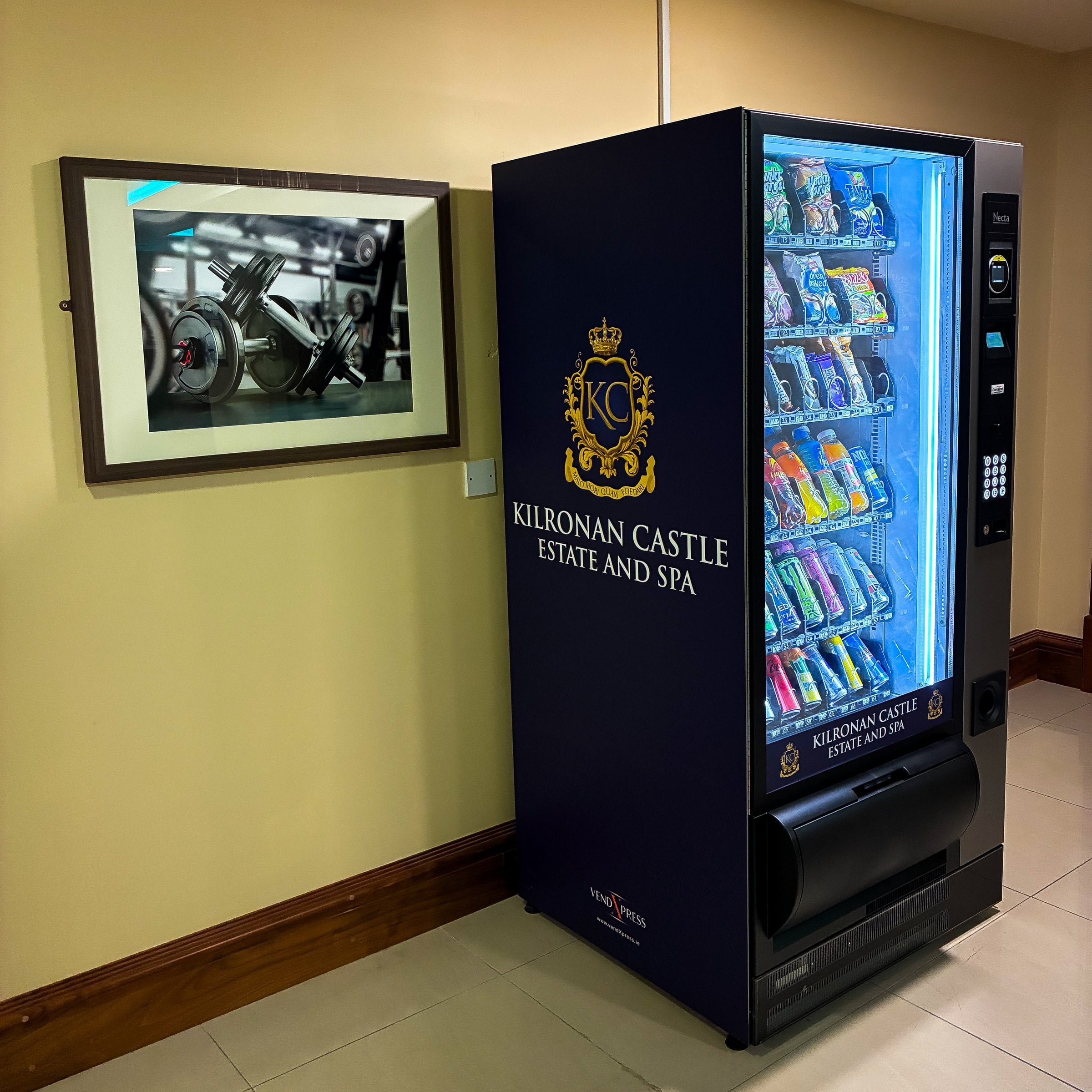 Necta snack and drinks vending machine in a navy Kilronan Castle Estate and Spa wrap, on site in a cream corridor beside a framed gym photograph