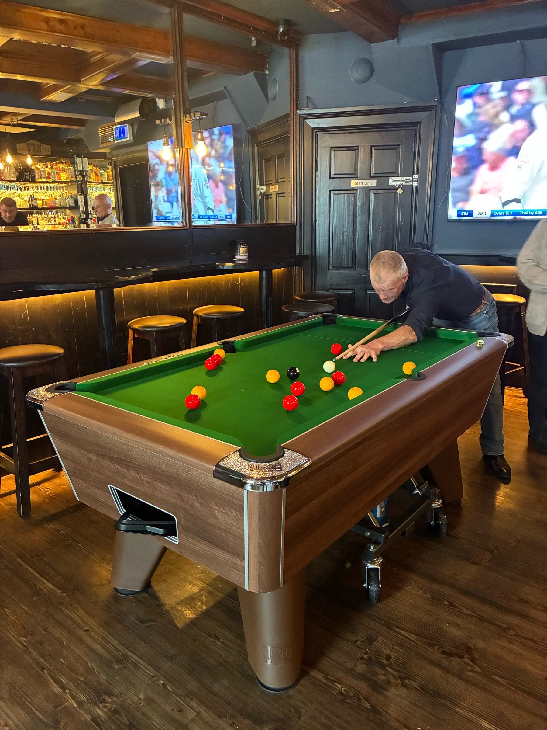 Supreme pool table with green cloth in a pub, a player lining up a shot, with the bar and a television behind